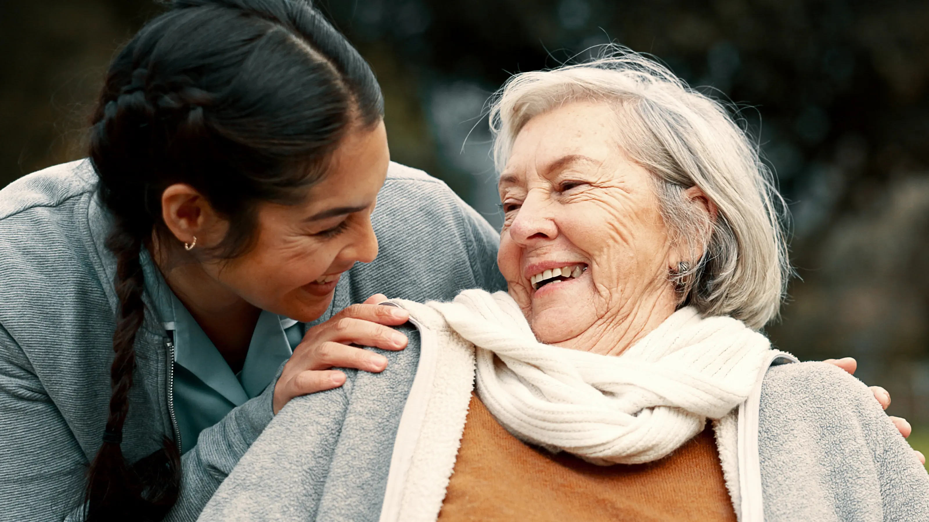 A young female Helper in a light blue uniform gently places her hand on an elderly woman's shoulder as they share warm smiles. Outdoors, they enjoy a moment of happiness together.