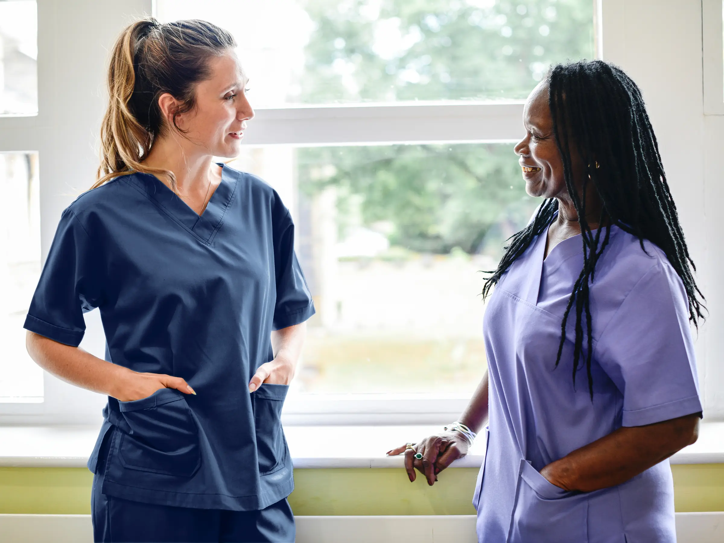 Two female healthcare professionals chat by a large window, sharing a warm, friendly moment. Natural light fills the room, highlighting the welcoming and professional atmosphere.